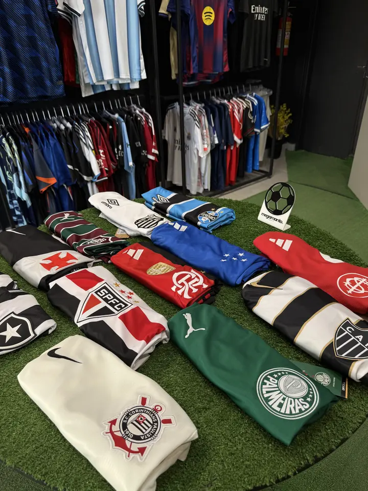 Camisas de Corinthians, Palmeiras, São Paulo, Flamengo, Cruzeiro e outros times dobradas sobre uma mesa de grama sintética